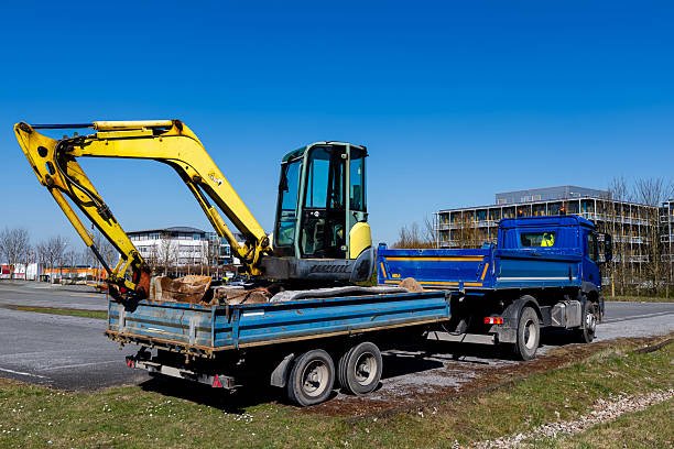 Small yellow excavator loaded onto a truck trailer. A truck is parked in a parking lot under a blue sky.